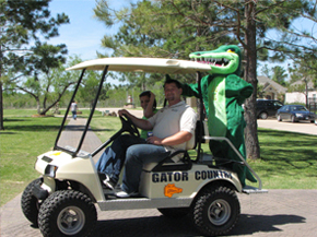 Two men in a golf cart with a person in an alligator costume on the back.